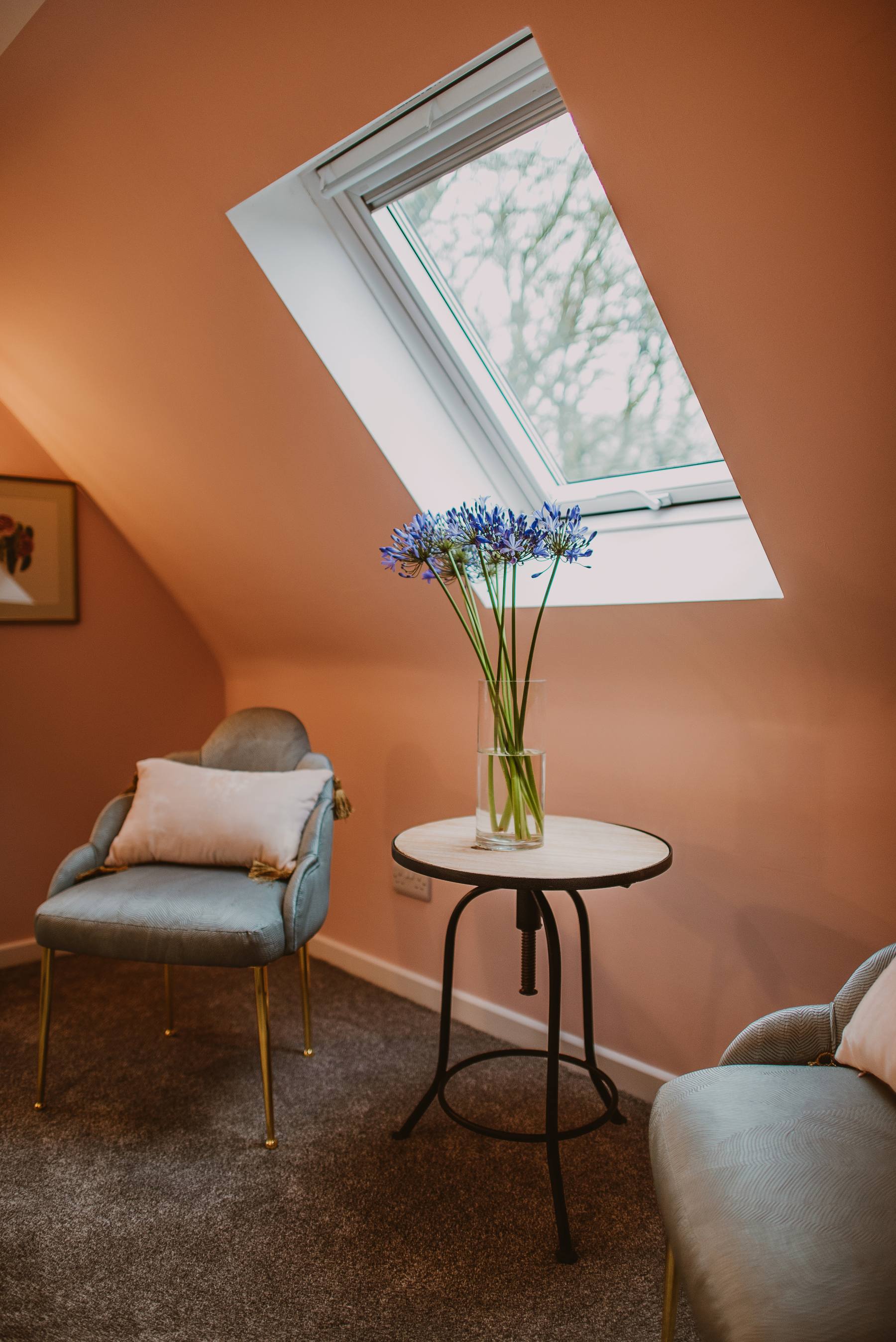 Two grey armchairs sit either side of a round table with a vase of agapanthus, a roof window lighting the sloped ceiling above.