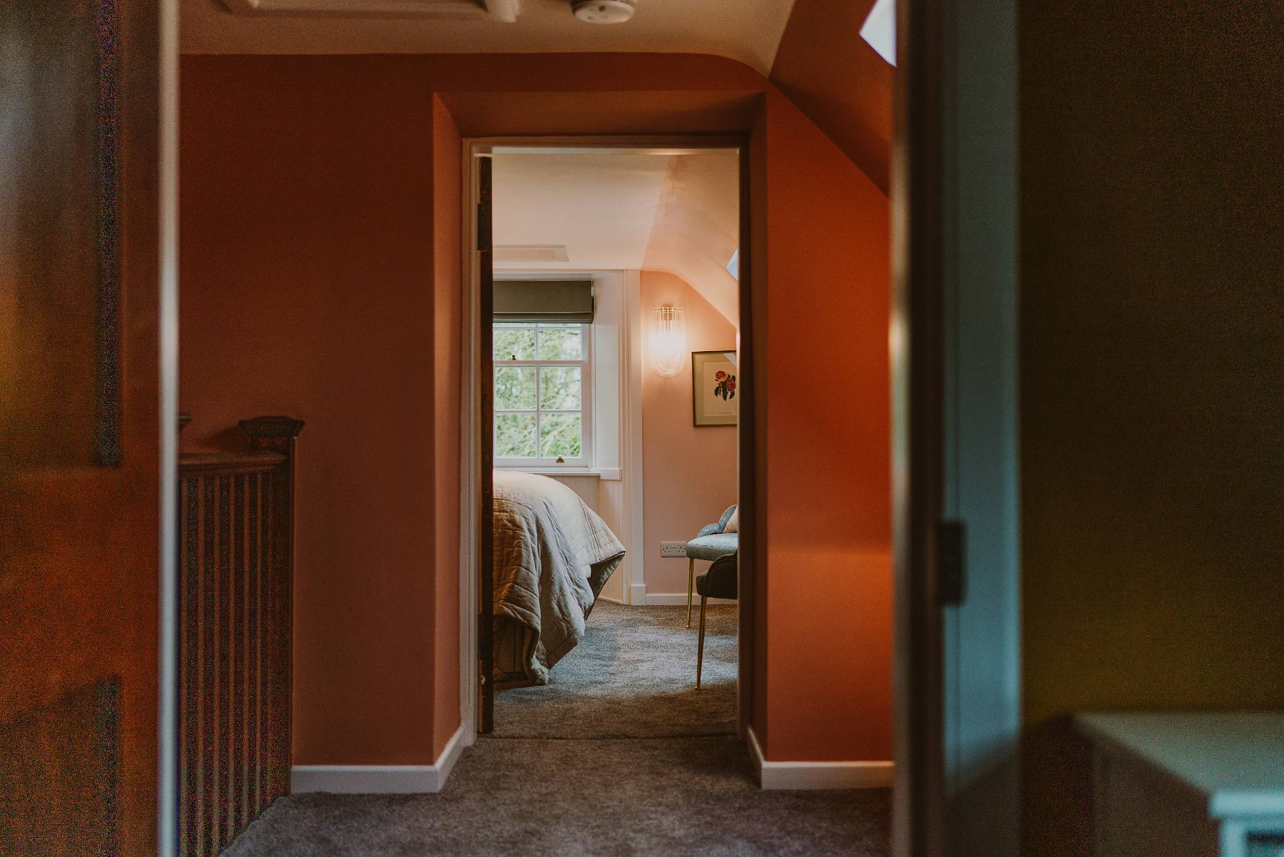 An open door leads into a terracotta-walled bedroom with a made bed and window seat, seen from a carpeted landing.