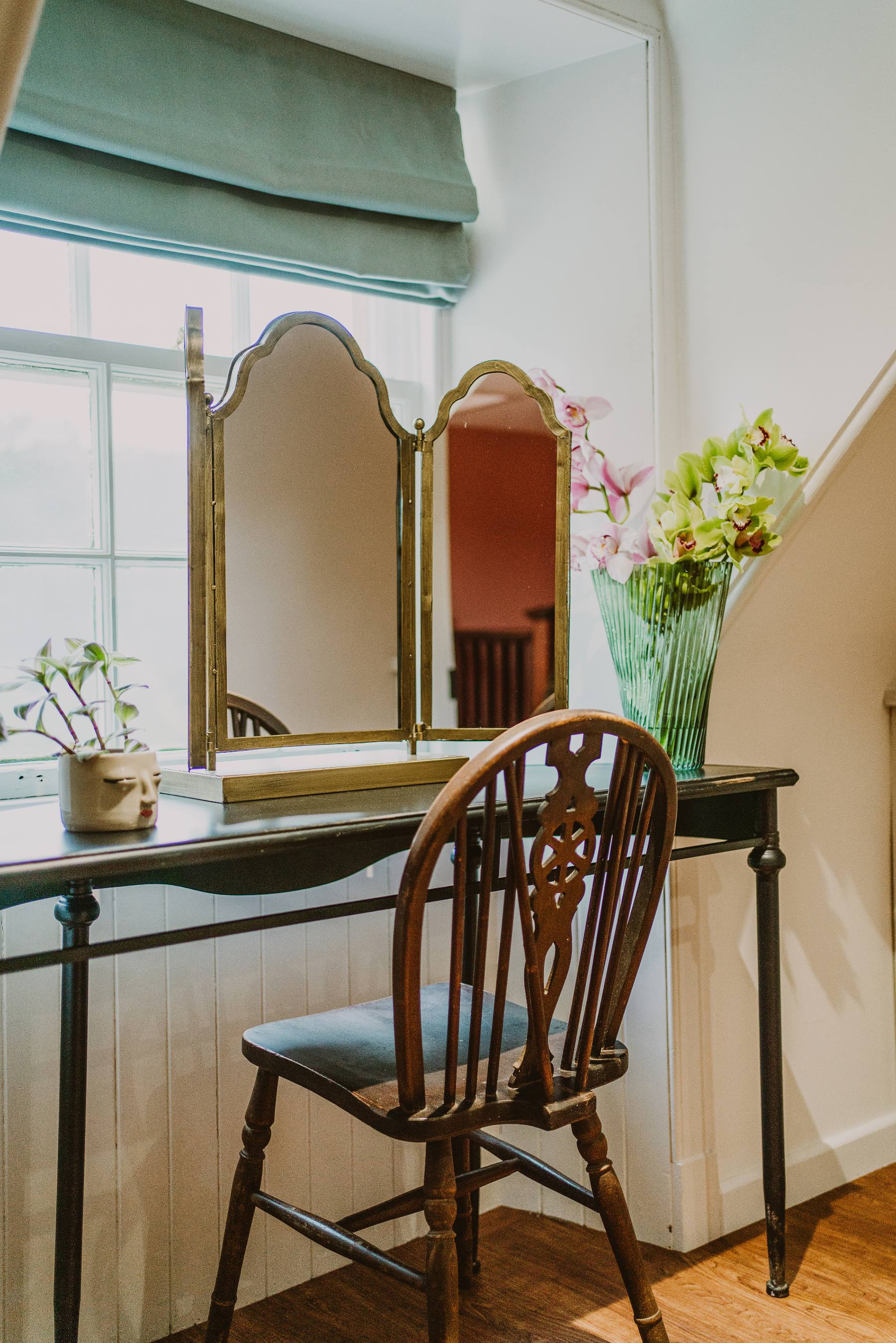A tri-fold mirror stands on a dressing table beside a vase of orchids, a wooden chair and staircase visible behind.