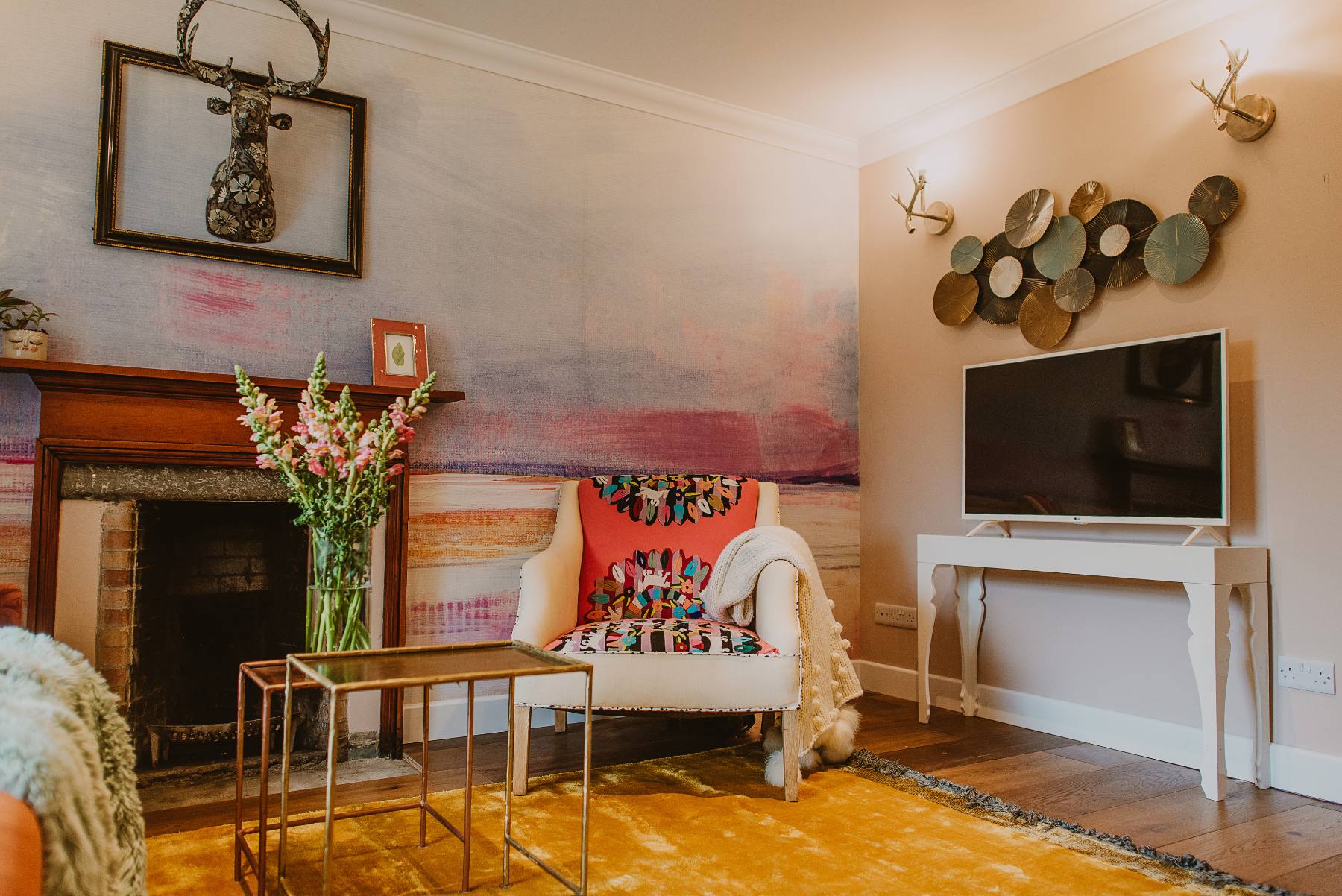 A mounted stag's head hangs above a tiled fireplace filled with flowers, a patterned armchair and a wall of decorative plates beside a mounted television.