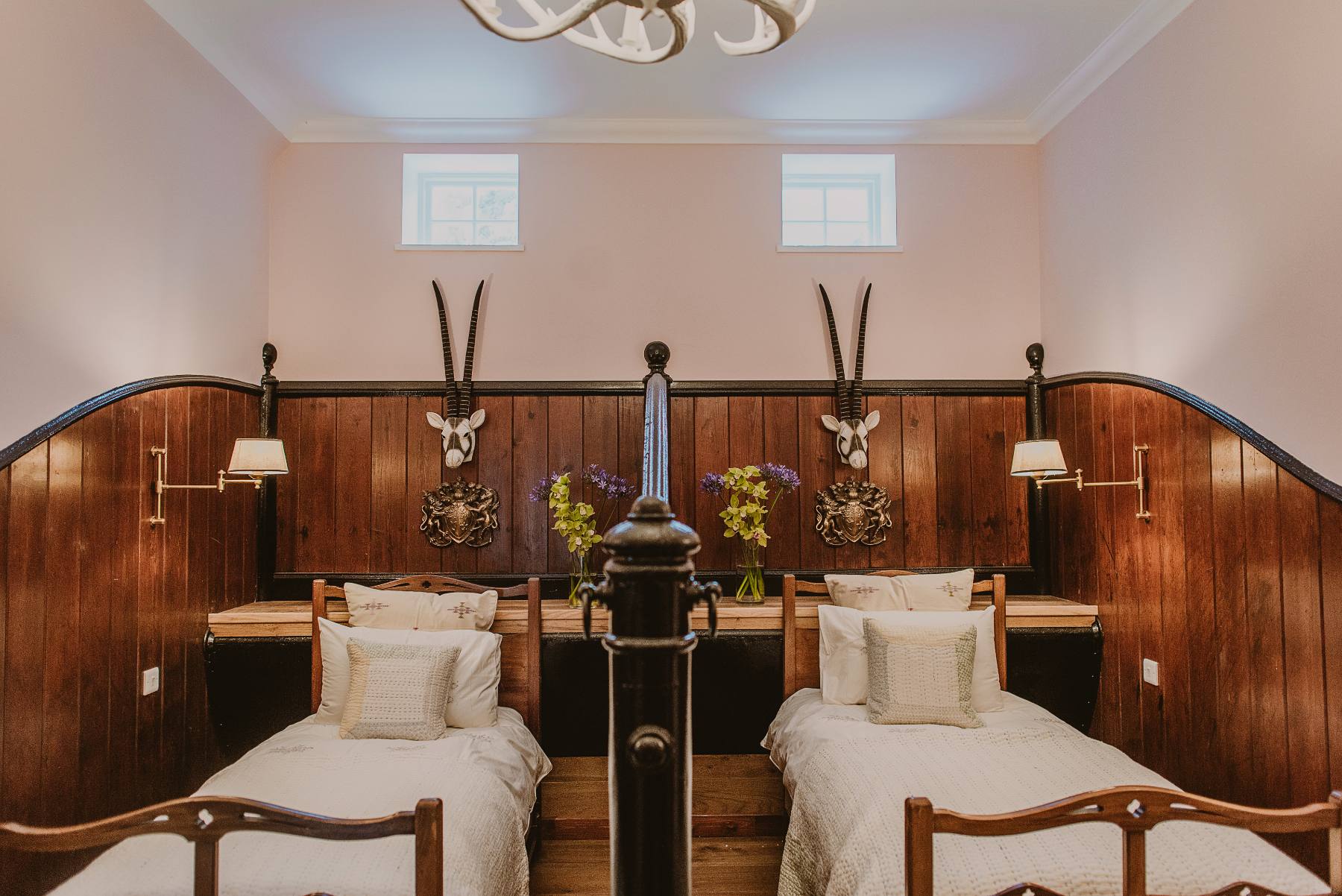 Twin beds with cream linen flank a dark panelled headboard hung with antlers, sconce lamps lit either side beneath a chandelier.