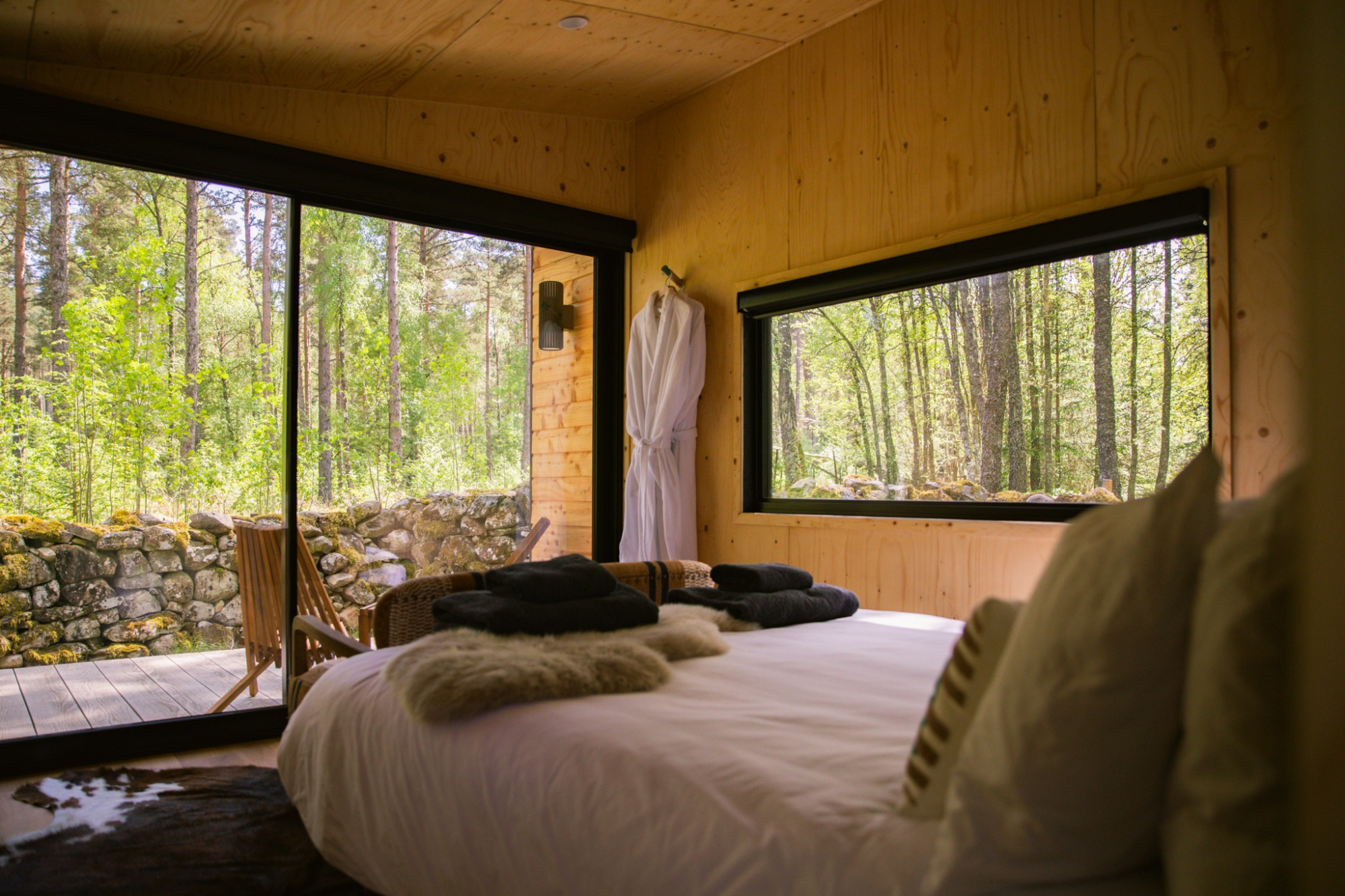 A bed with white linen and a sheepskin throw sits before floor-to-ceiling windows looking onto woodland, a wicker chair on the deck outside.