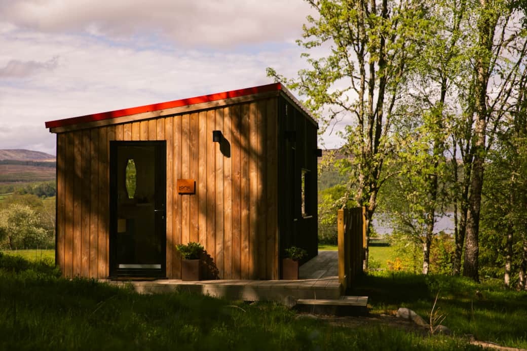 A timber cabin with a red roofline and glazed door stands among trees, potted plants either side of the entrance.