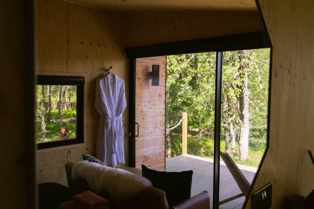 A white robe hangs on a hook beside a glazed door opening onto a timber deck surrounded by trees, a sofa in the foreground of the panelled room.