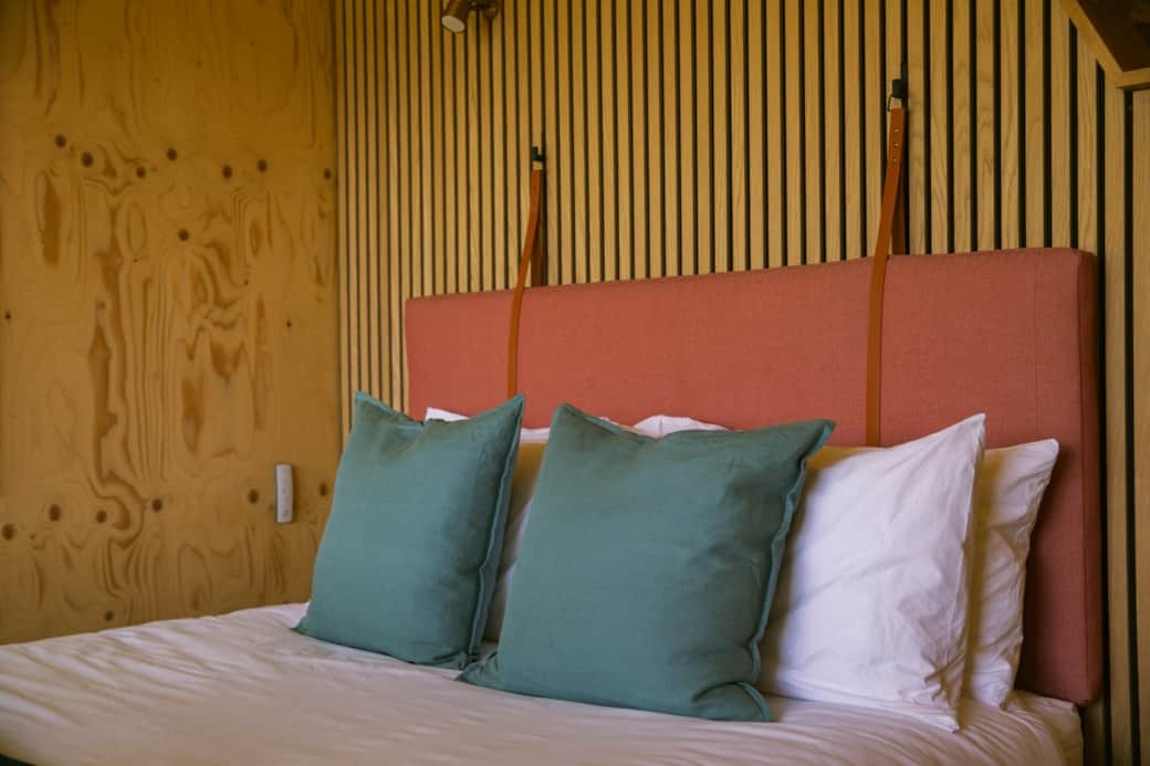 A bed with white linen and teal cushions sits before a coral upholstered headboard on a slatted timber wall.