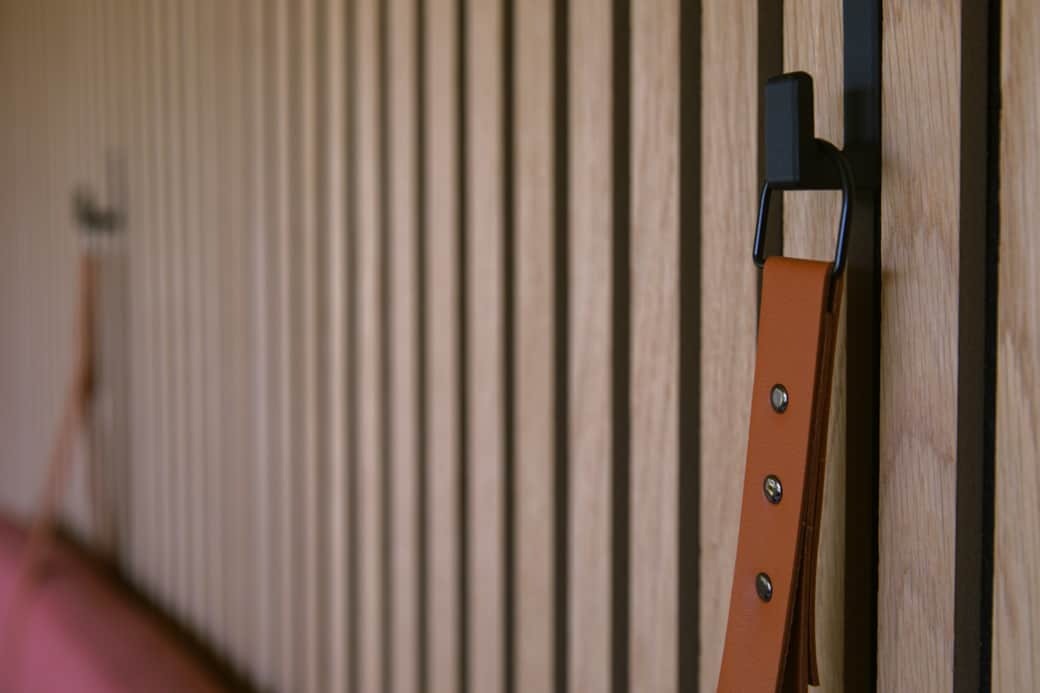 A leather strap hook is mounted on a slatted timber wall, a pink headboard blurred in the background.