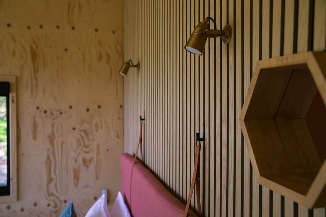 A pink upholstered headboard sits against a slatted timber wall fitted with two brass reading lamps and leather strap hooks.