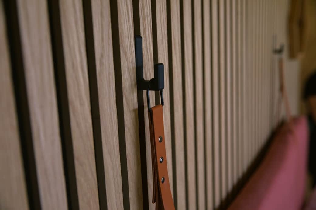A leather strap hook is mounted on a slatted timber wall beside a pink headboard, a light fixture visible in the background.
