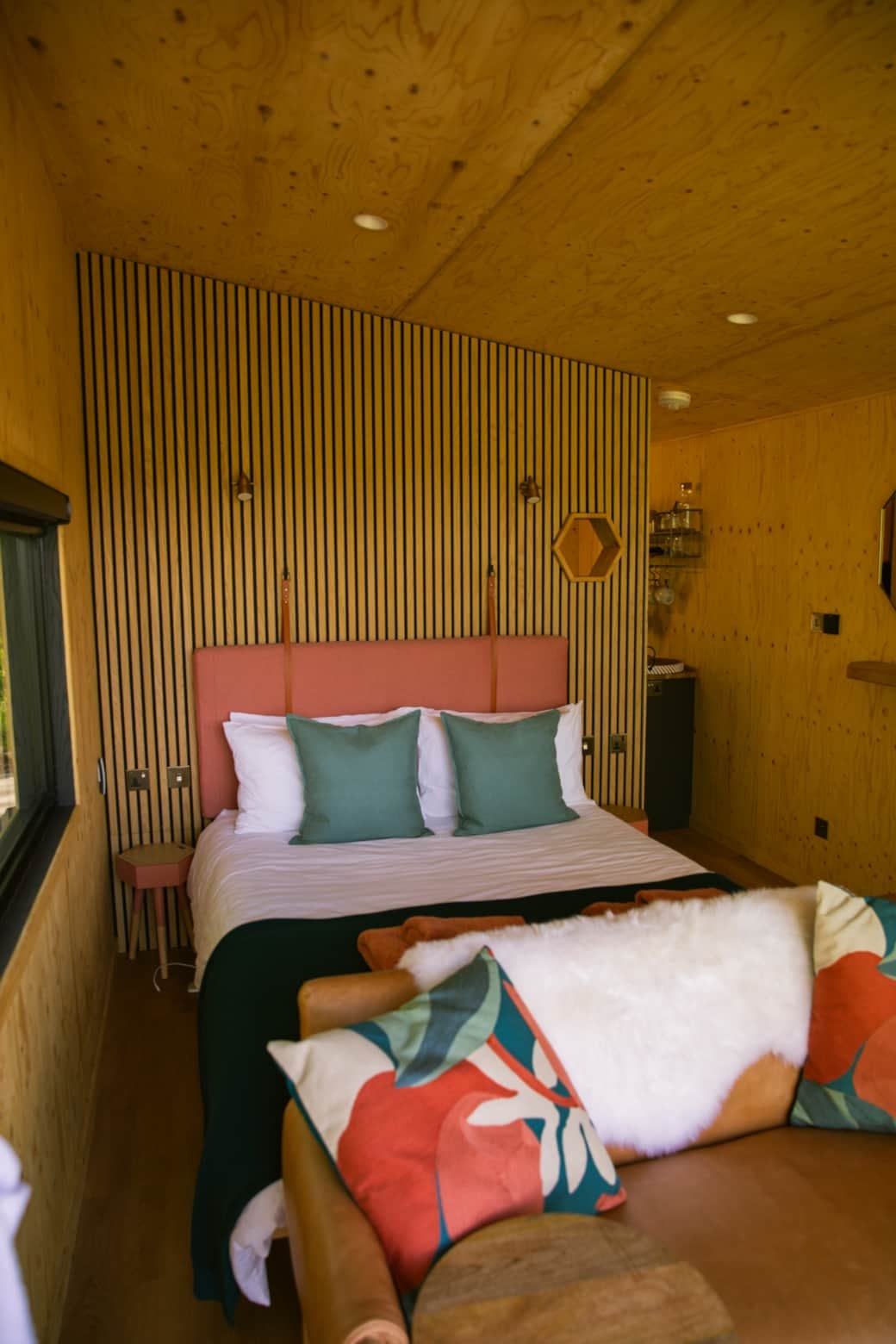 A bed with a coral upholstered headboard and teal cushions sits below a hexagonal mirror, a leather sofa with patterned cushions in the foreground.