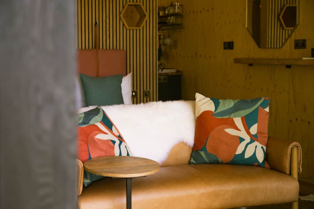 A tan leather sofa holds a white sheepskin and patterned orange cushions, a hexagonal mirror and shelving on the panelled wall behind.