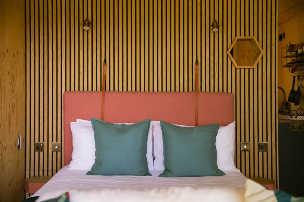 A bed with white linen and teal cushions sits before a coral upholstered headboard on a slatted timber wall, a hexagonal shelf to the right.