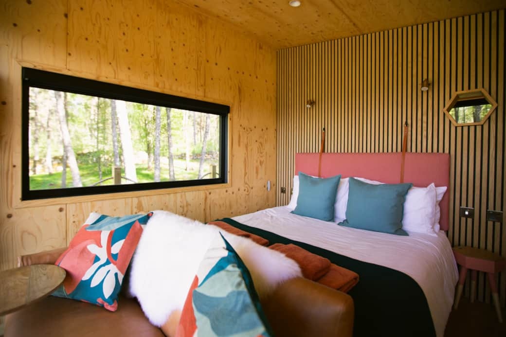 A bed with a coral headboard and teal cushions sits beside a large window looking onto woodland, a leather sofa with patterned cushions in the foreground.