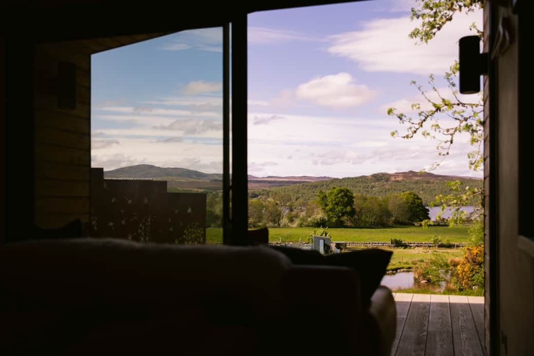 A loch and hills are framed by an open glazed door, a sofa in the foreground of a timber deck.