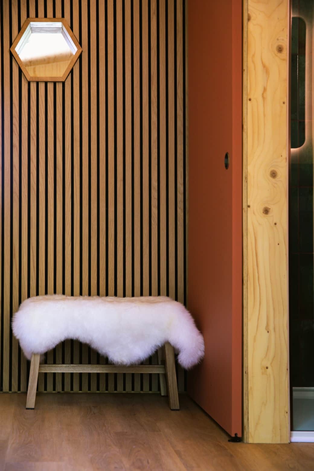A sheepskin-covered bench sits below a hexagonal mirror on a slatted timber wall, a coral door leading to a shower room beside it.