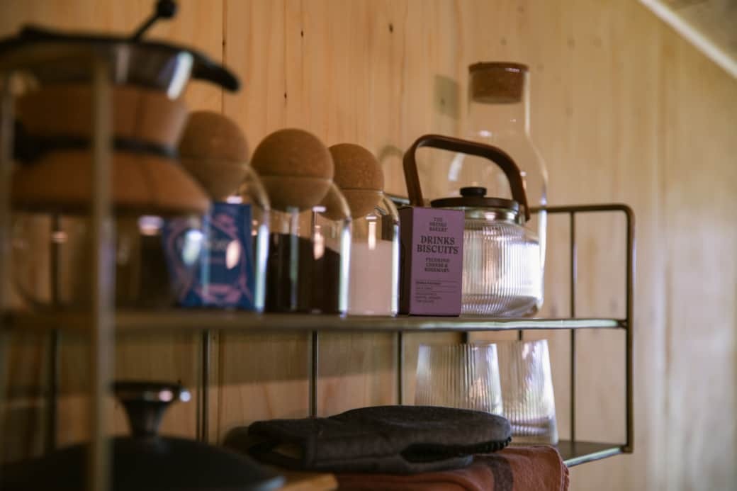 A kettle, glasses and a box of biscuits are arranged on an open shelving unit against a timber-panelled wall.