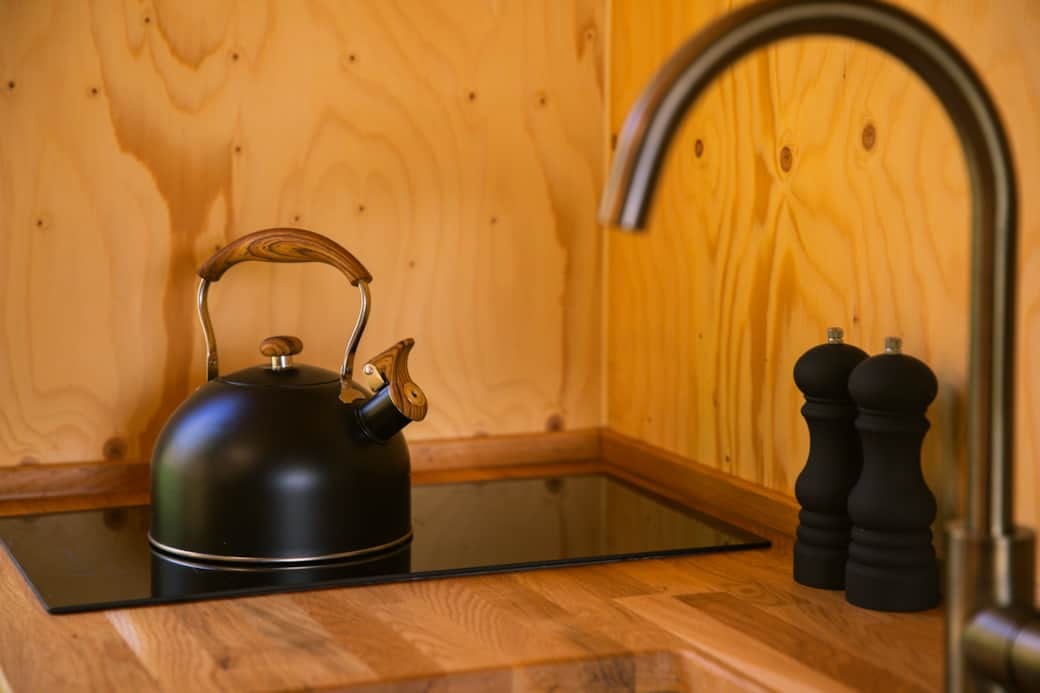 A black kettle and two pepper grinders stand on a wooden counter beside a tap, set against a plywood wall.