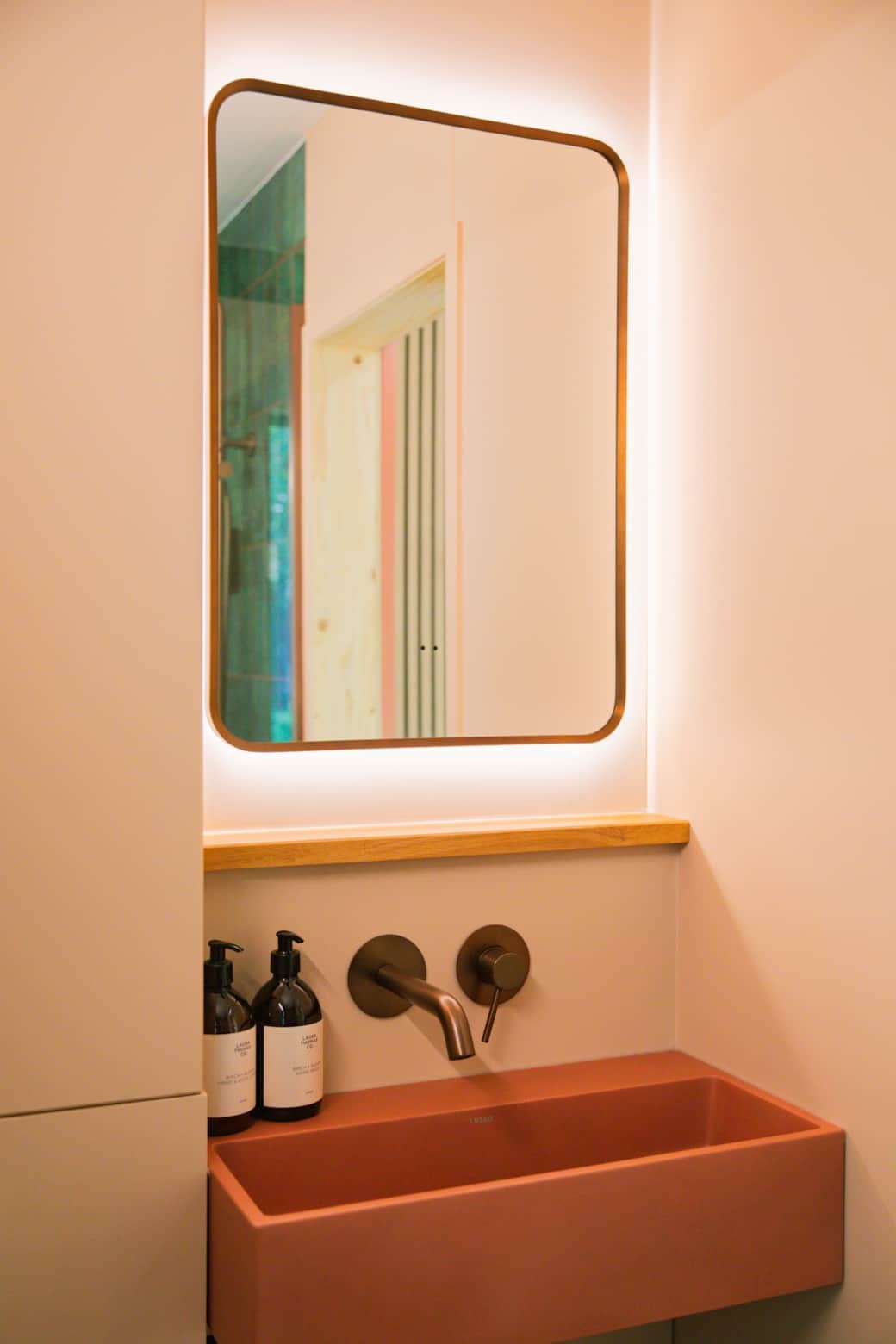 A wall-mounted mirror with a warm backlit glow hangs above a pink sink, two amber pump bottles set into the wall-mounted taps.