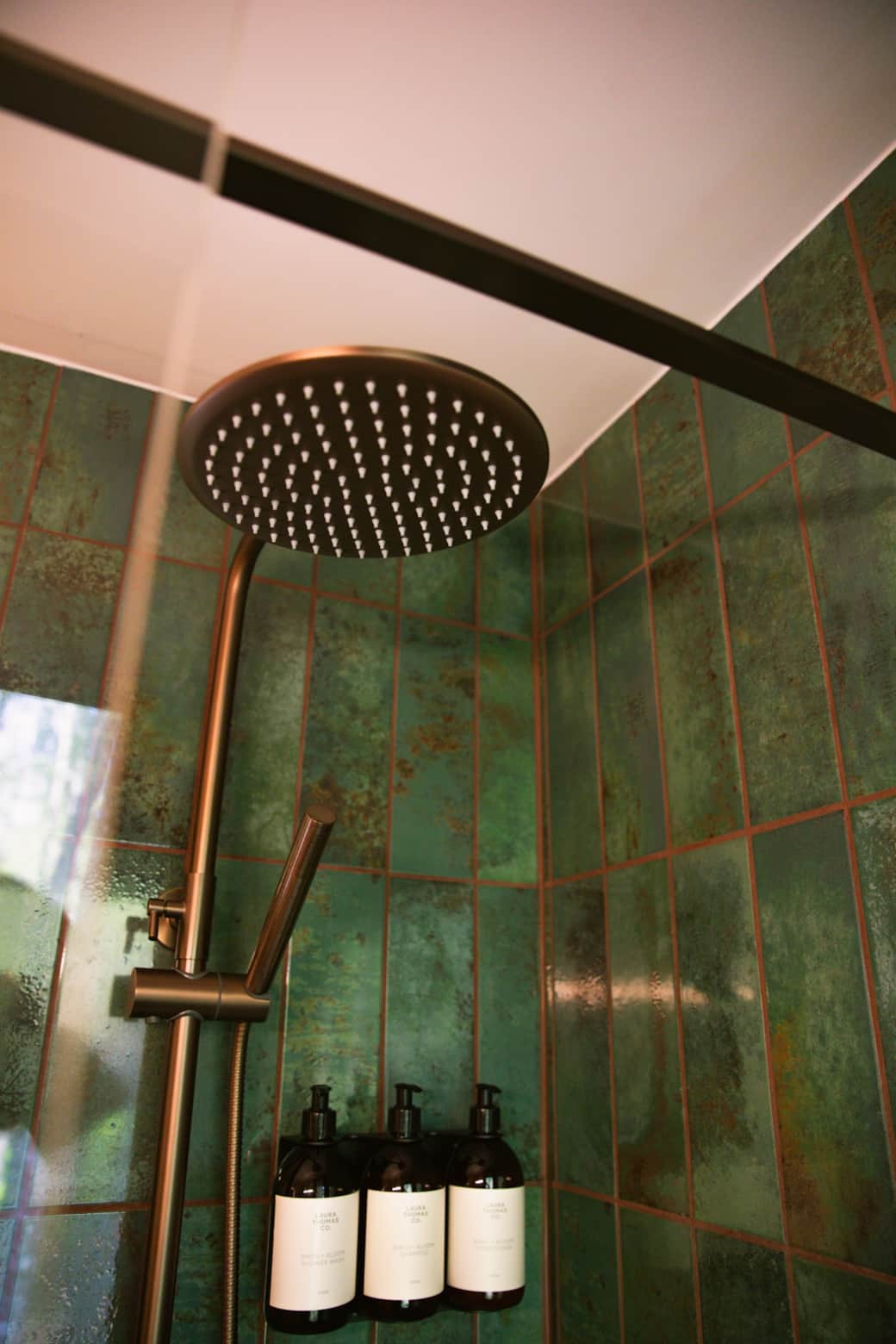 A rainfall shower head and handheld attachment are mounted in a green tiled shower, three amber toiletry bottles on the wall below.