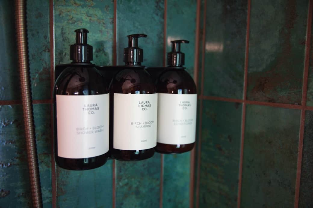 Three amber pump bottles of toiletries hang from a rail against a teal tiled shower wall.