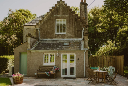 The stone cottage seen from the garden, with crow-stepped gables, a wooden bench and a hot tub set on the patio to the right.