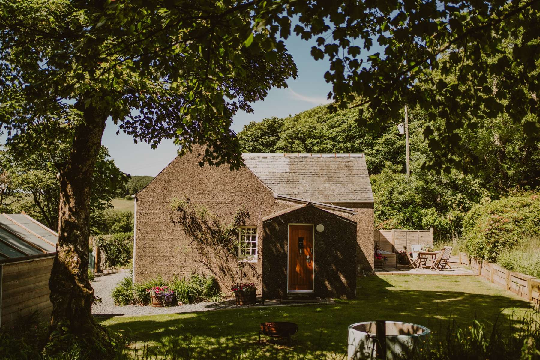 A stone cottage with a slated roof sits shaded by overhanging trees, an orange door and picnic table set on the lawn beyond a gravel path.
