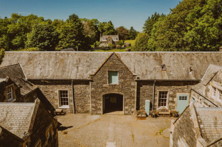 An aerial view shows a stone courtyard building with a gabled archway, other roofs and trees surrounding it.