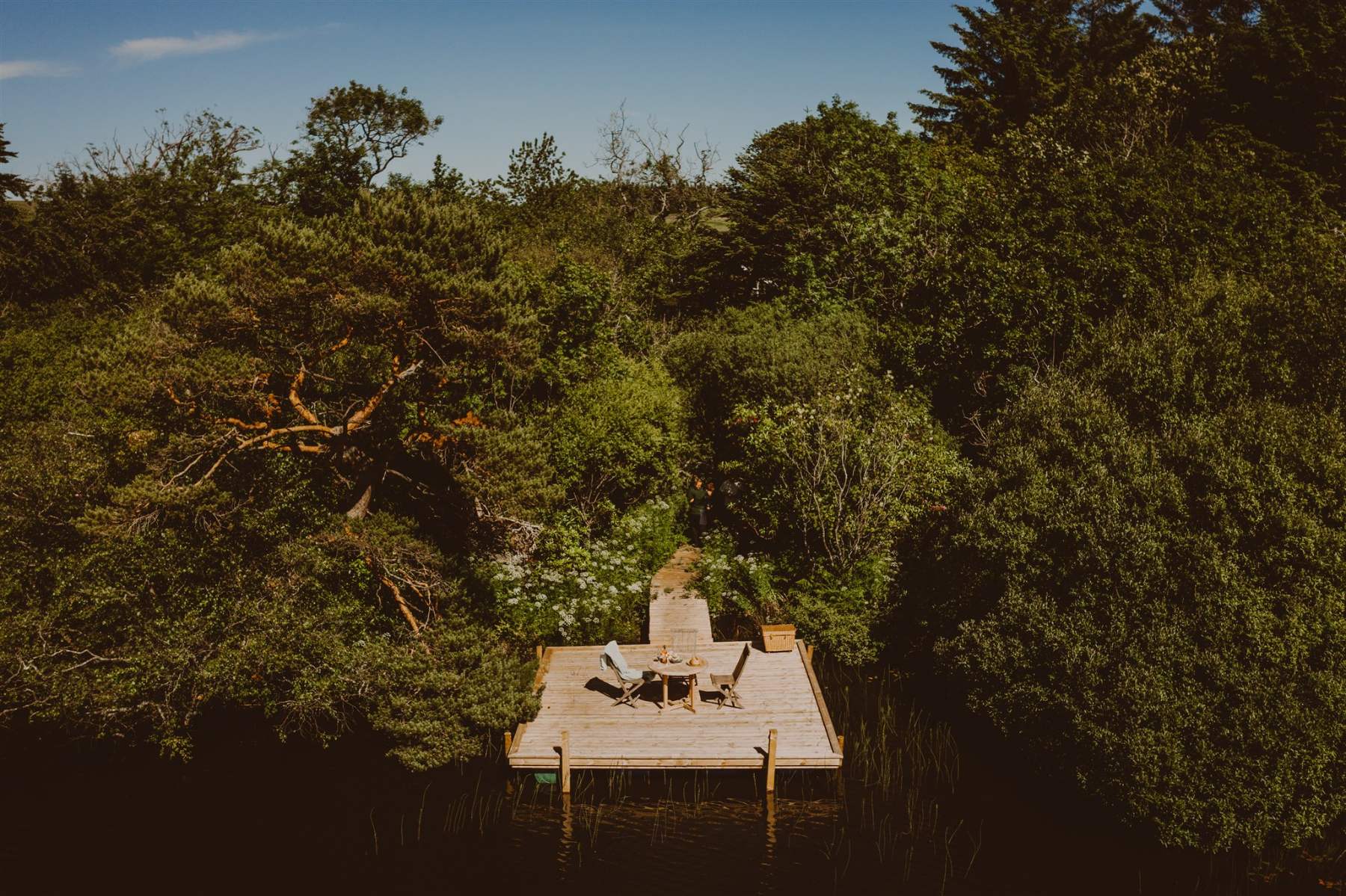 A wooden jetty with chairs extends into a loch framed by trees, a gnarled branch overhanging the water.