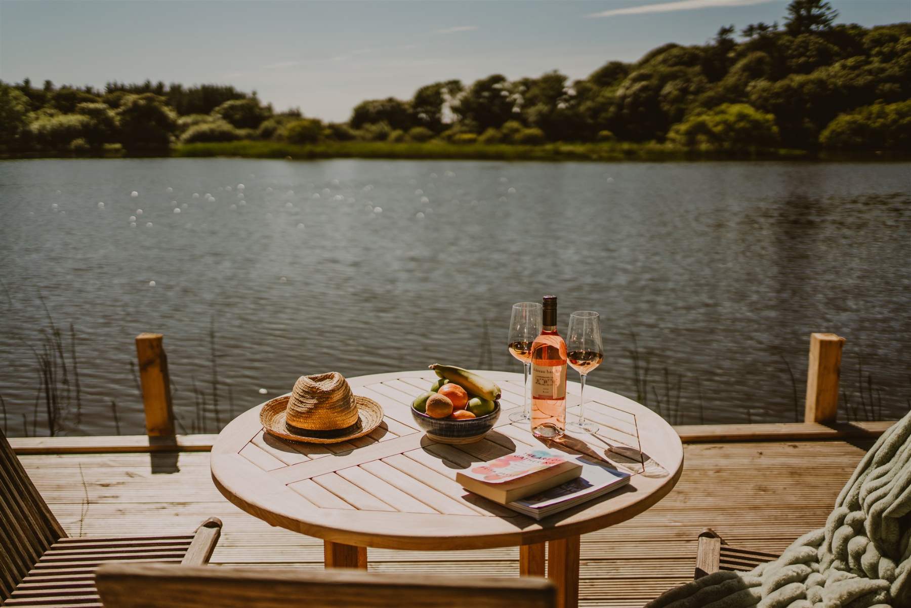 A straw hat, a bowl of fruit and a bottle of rosé with two glasses sit on a round wooden table beside a loch, a book resting nearby.