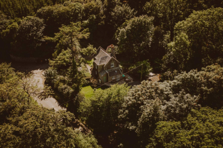 An aerial view shows a turreted stone house nestled among dense trees beside a curving driveway.