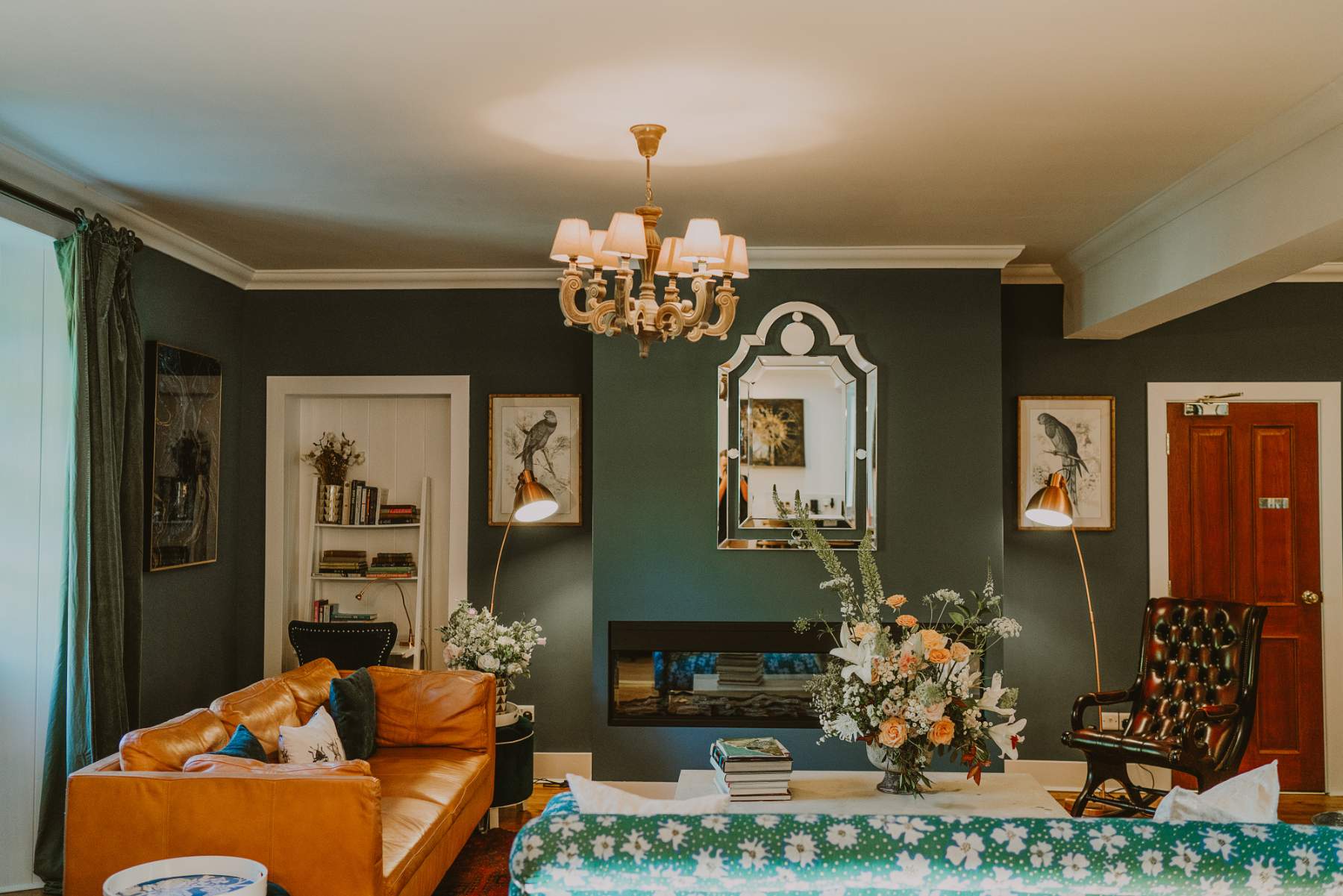 A chandelier hangs above a dark green sitting room with a leather sofa and a floral sofa around a fireplace, a gilt mirror on the wall.
