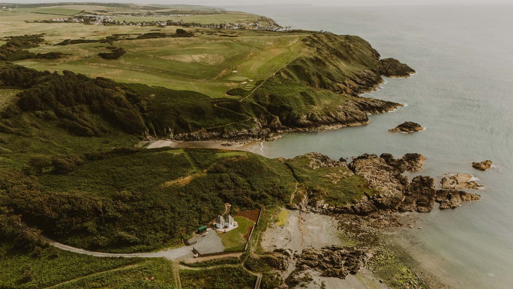 An aerial view shows a rocky headland and golf course above a sandy cove, a white building nestled among trees near the shore.