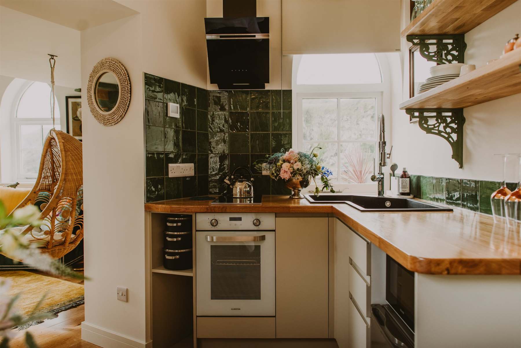 A galley kitchen with green tiled splashback and a wooden hanging egg chair visible beyond, a vase of flowers on the counter beside the oven.