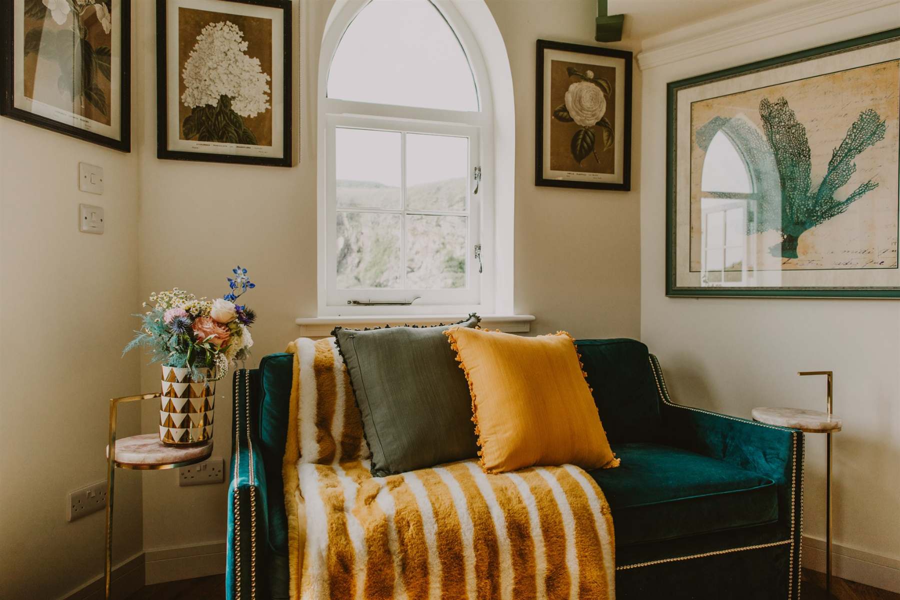 A teal velvet sofa with mustard and olive cushions sits below an arched window, framed botanical prints and a striped throw completing the corner.