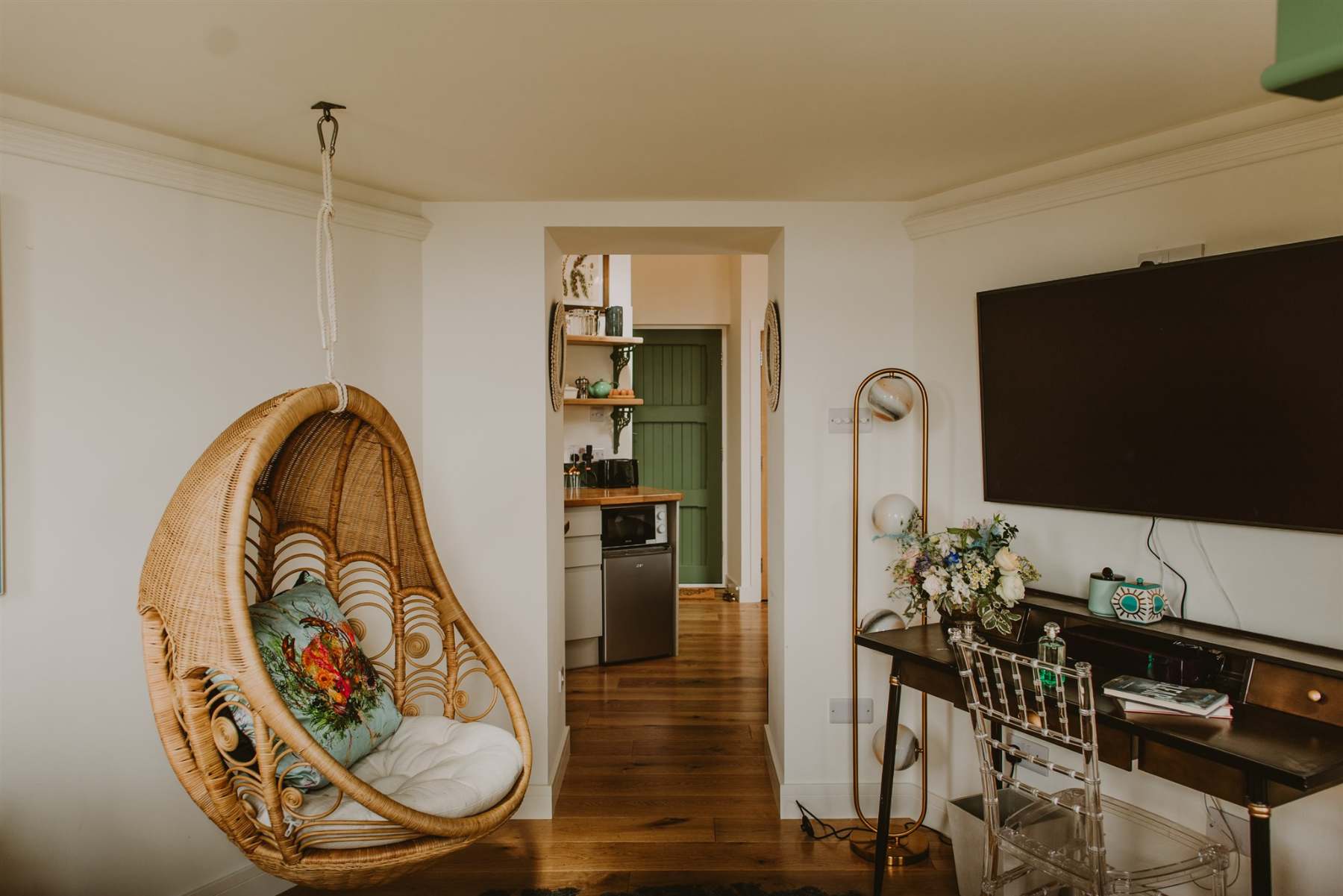 A wicker hanging egg chair with a cushion sits in a corner, a mounted television and bar cart visible beside a hallway leading to a kitchen with green cabinetry.