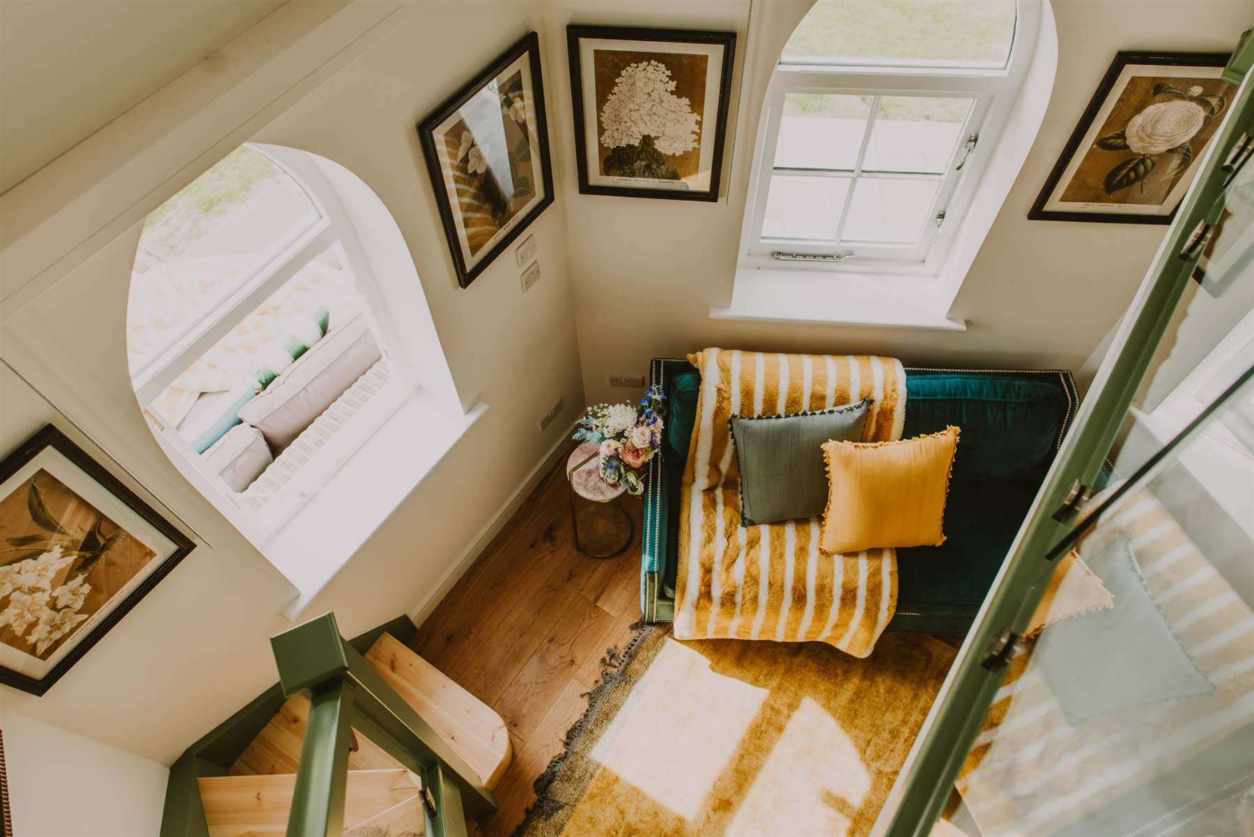 A mezzanine landing looks down on a teal sofa with striped throw, framed botanical prints on the white walls around two arched windows.