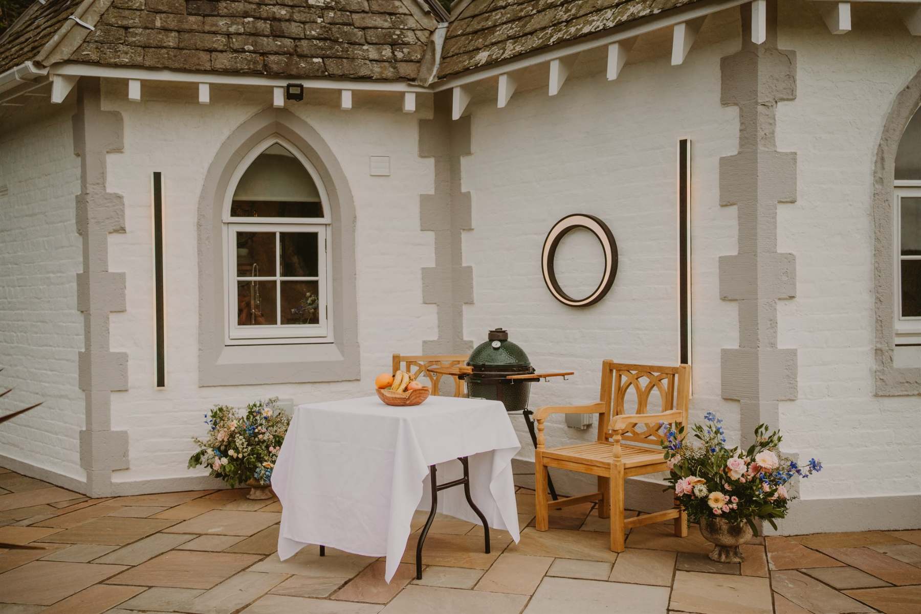 A round table draped in white cloth stands beside a green ceramic barbecue on a paved patio, wooden chairs and urns of flowers either side of an arched window.
