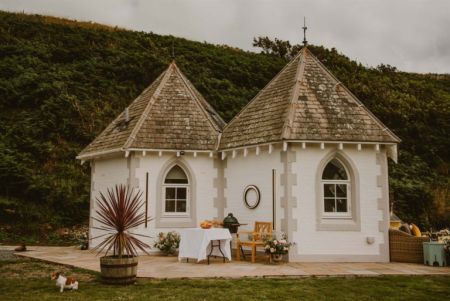 A white gatehouse with twin turreted roofs stands on a lawn, a round table set for tea beside a green ceramic barbecue and potted palm.