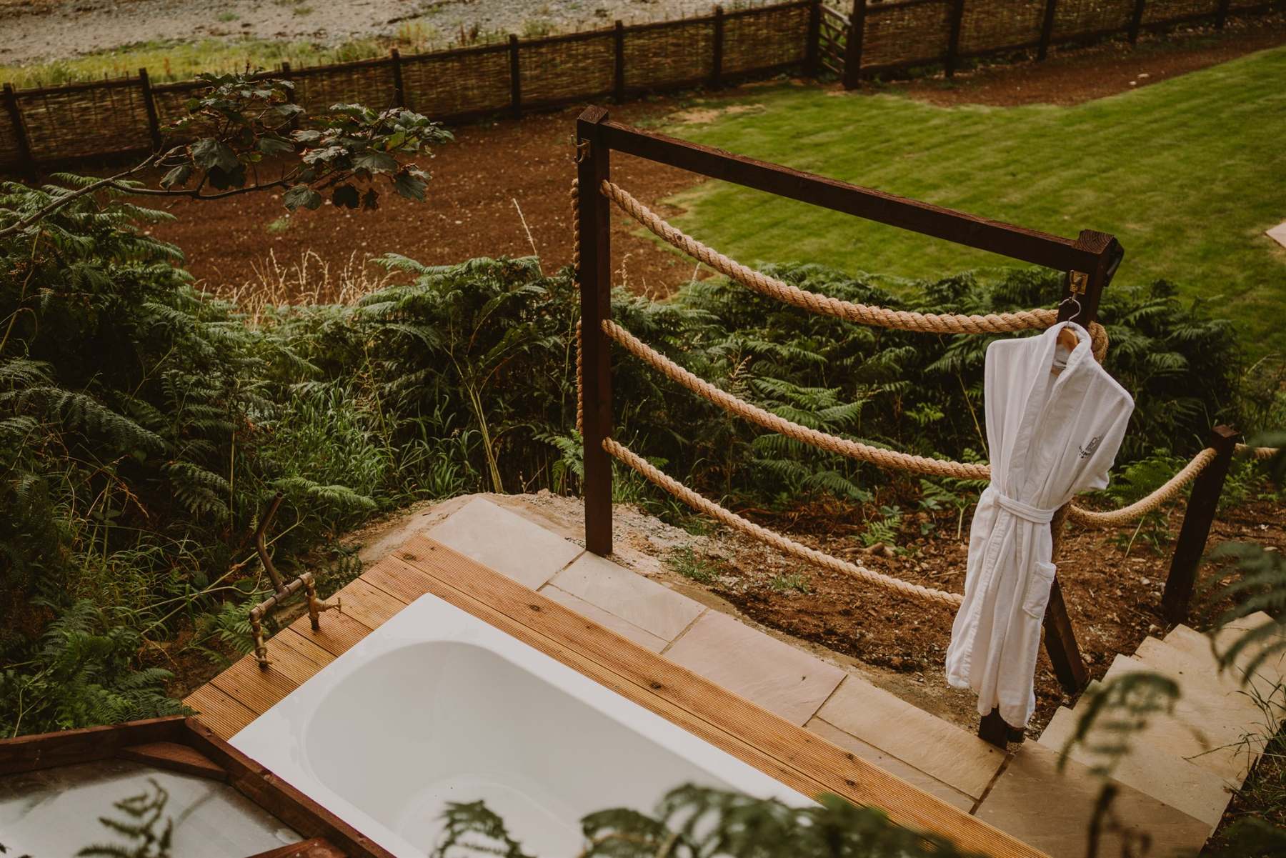 A white robe hangs on a rope railing beside a freestanding bath on a timber deck, ferns and a lawn visible below.