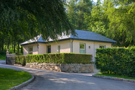 A single-storey lodge with a slated roof sits behind a stone wall and clipped hedge, mature trees shading the curving drive.