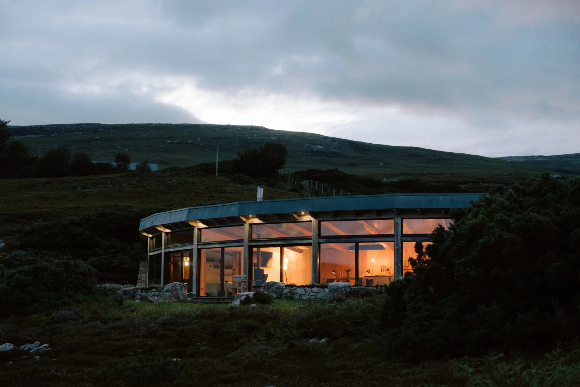 A curved timber house glows with lit windows at dusk, set low into a grassy hillside.